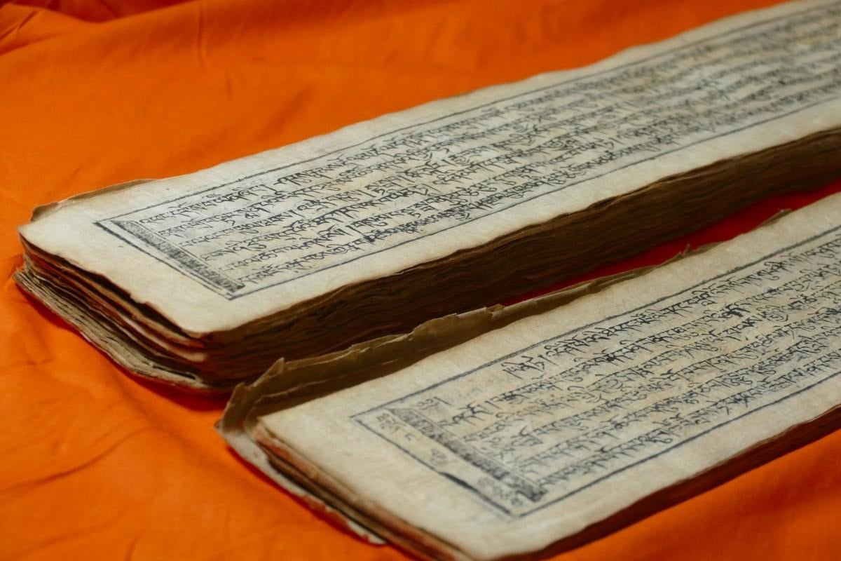 Zwei lange, historische Manuskriptbände mit handgeschriebenem Text liegen auf einem orangefarbenen Stoff. Die Seiten wirken gealtert und zeigen eine traditionelle Schrift, vermutlich aus dem tibetischen Kulturraum.