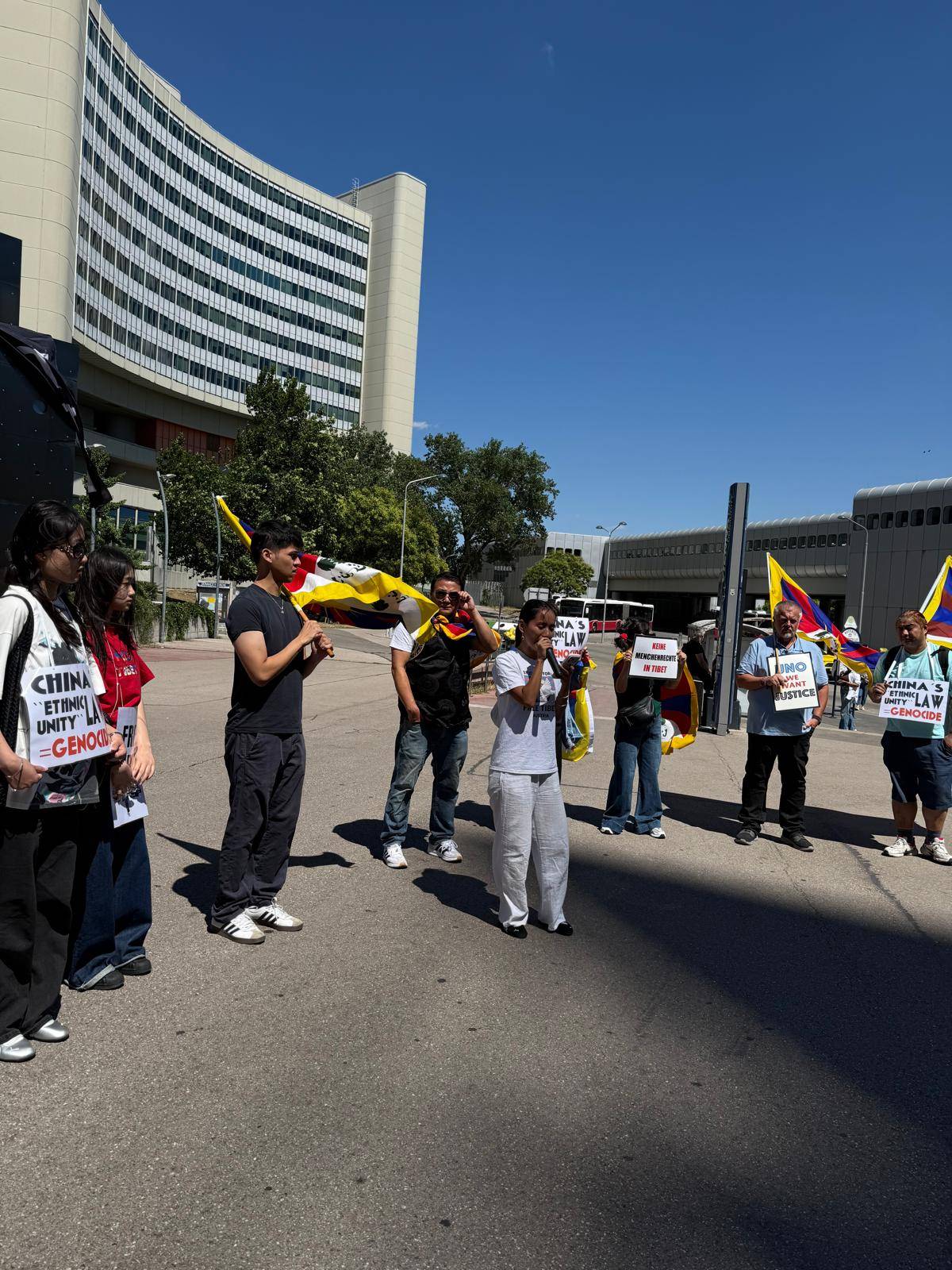 Vor dem Vienna International Centre stehen mehrere Menschen mit Tibet-Flaggen und Protestschildern in einem Halbkreis. Eine Person spricht mit einem Mikrofon zu den Teilnehmenden. Auf Schildern steht unter anderem „China's Ethnic Unity Law = Genocide“, „Keine Menschenrechte in Tibet“ und „UNO We Want Justice“. Im Hintergrund sind das UNO-Gebäude und ein wolkenloser Himmel zu sehen.
