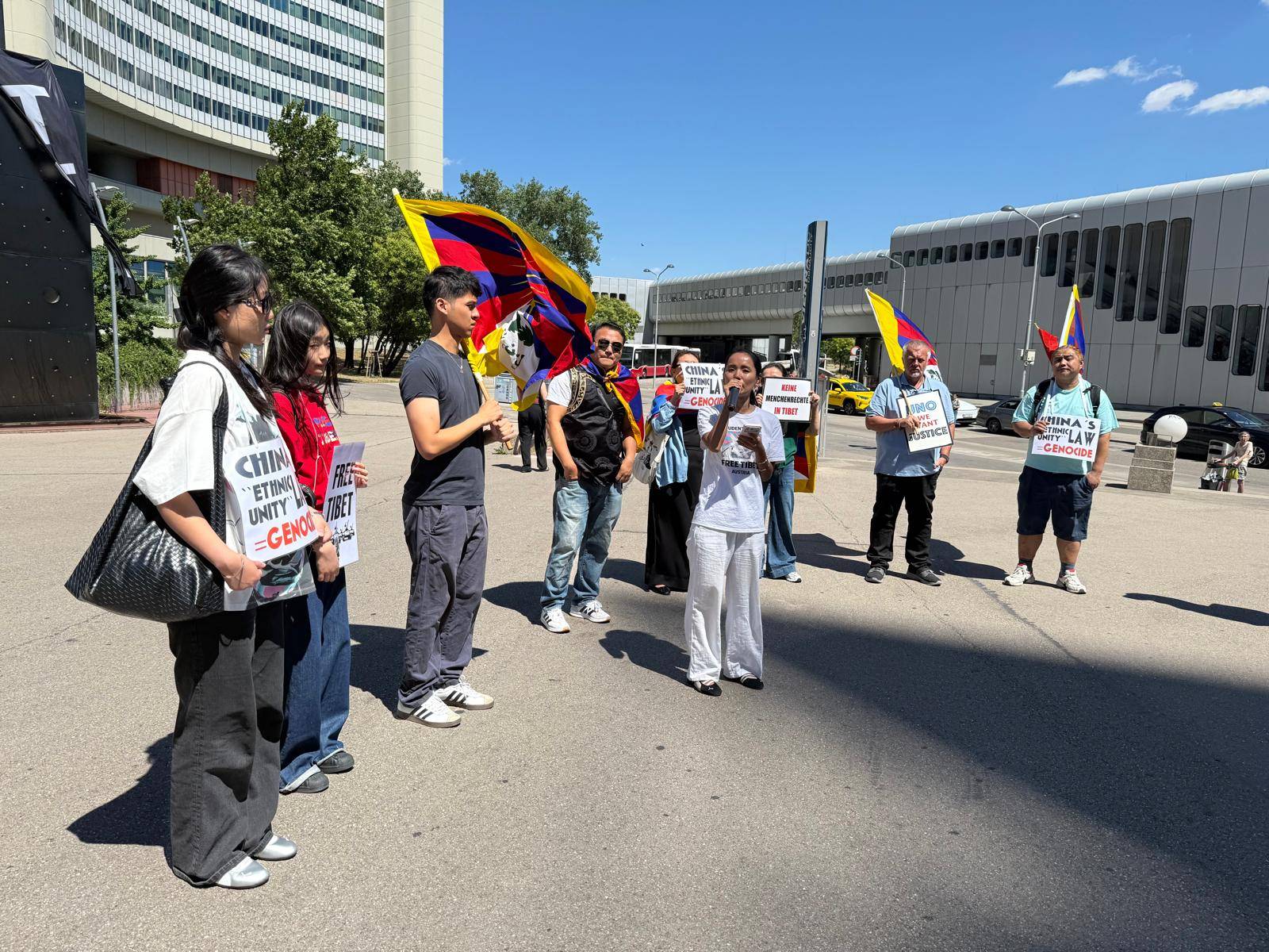 Eine Person spricht vor einer kleinen Kundgebung vor dem UNO-Gebäude in Wien. Die Teilnehmenden halten Tibet-Flaggen und Schilder mit Forderungen nach Menschenrechten und Gerechtigkeit für Tibet. Die Gruppe steht auf einem Platz in der Mittagssonne vor dem Vienna International Centre.