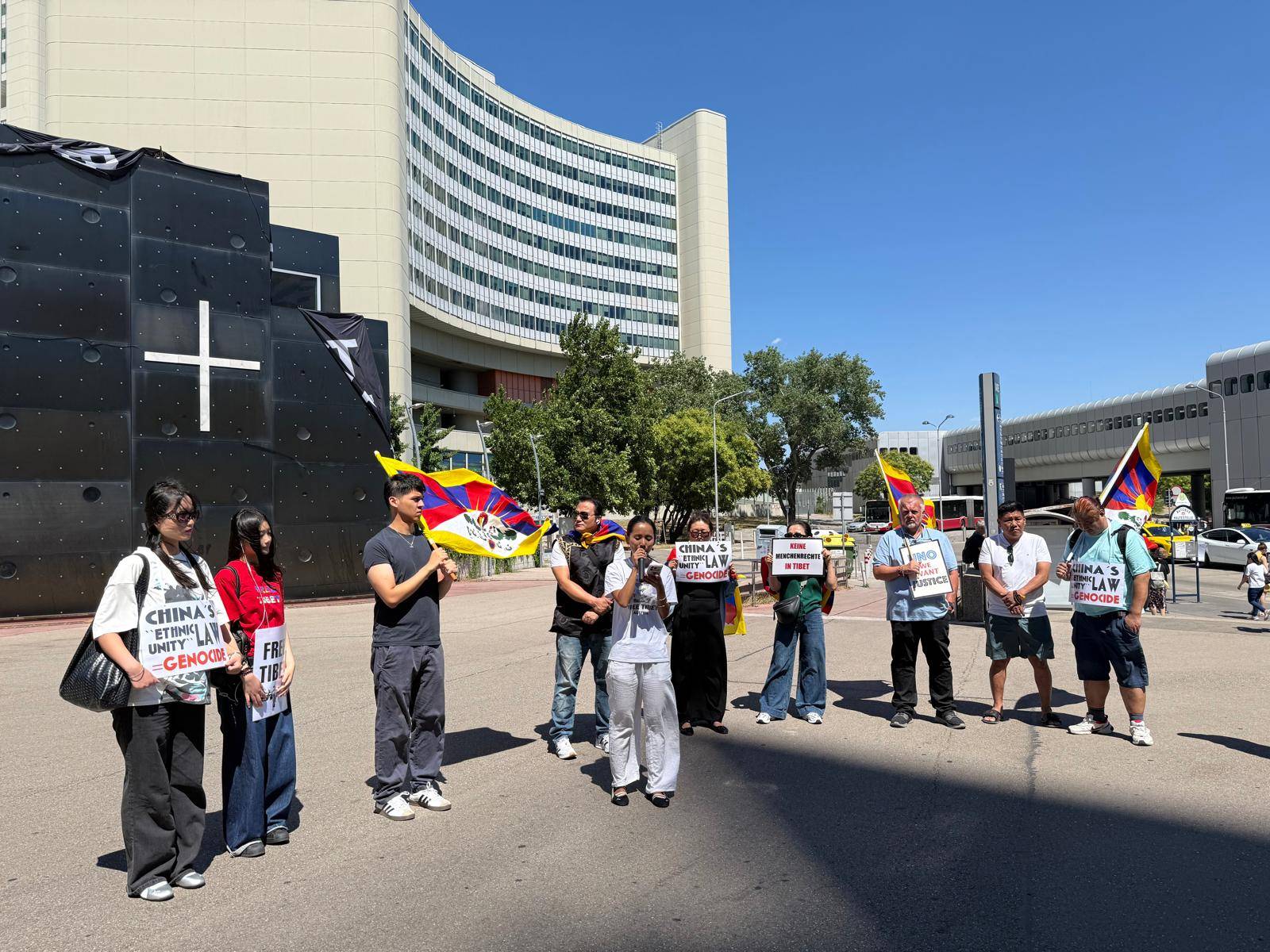 Weitwinkelaufnahme einer Kundgebung vor dem Vienna International Centre. Rund zehn Menschen stehen mit Tibet-Flaggen und Protestschildern zusammen, während eine Person eine Rede hält. Im Hintergrund sind die markante Fassade des UNO-Gebäudes und weitere Gebäude des UNO-City-Areals zu sehen.