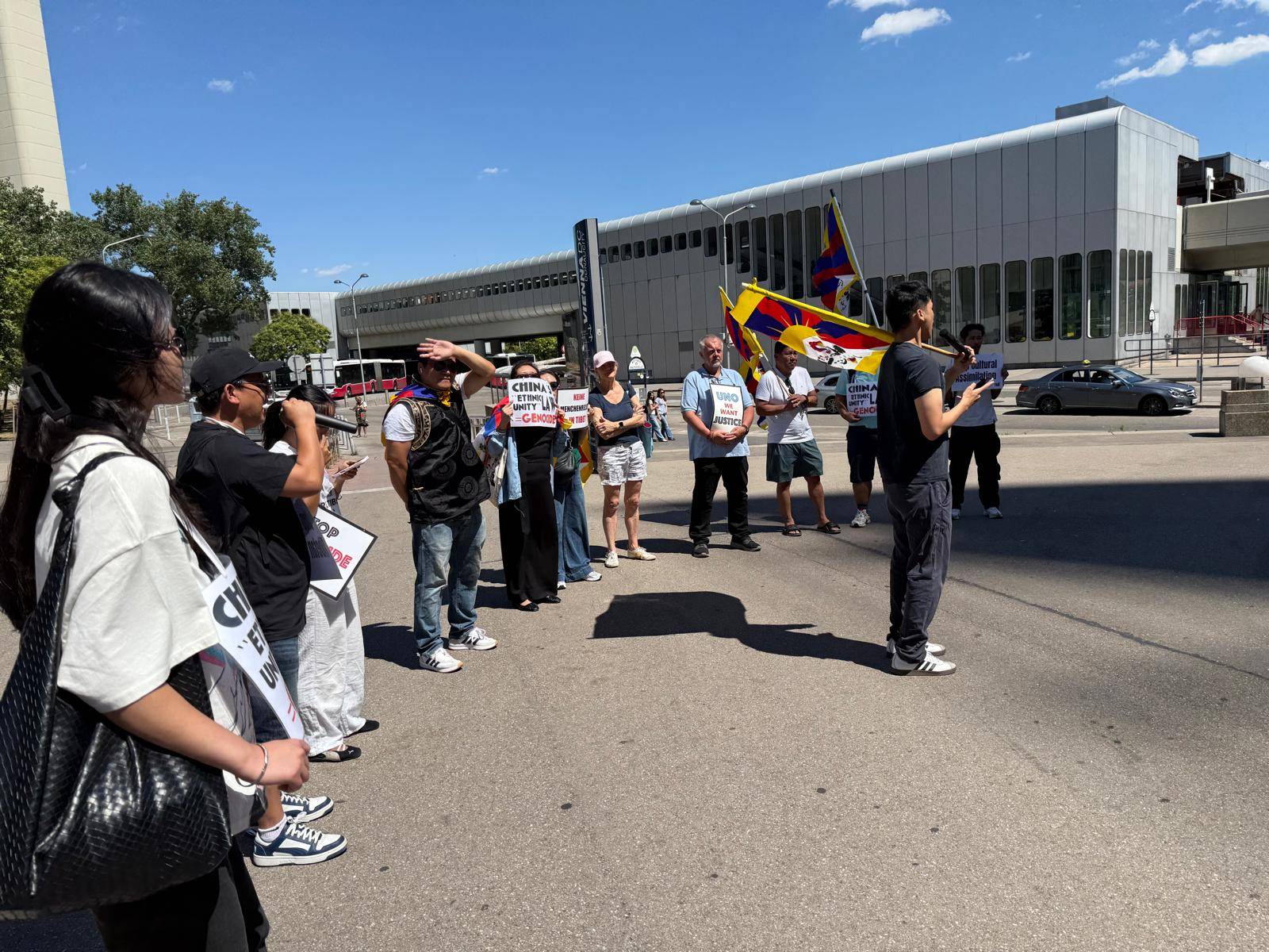 Seitliche Aufnahme der Kundgebung vor dem UNO-Gebäude. Eine Person spricht mit einem Mikrofon zur Gruppe. Mehrere Teilnehmende tragen Tibet-Flaggen oder halten Schilder mit Aufschriften wie „China's Ethnic Unity Law = Genocide“ und „UNO We Want Justice“. Eine große Tibet-Flagge weht im Wind.